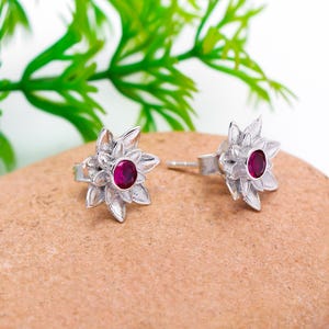 Könnte beinhalten: Ein Paar silberfarbene Ohrstecker. Jeder Ohrring hat ein Blumenmuster mit mehreren Blütenblättern, die einen runden, roten Edelstein umgeben. Die Ohrringe werden auf einer braunen, strukturierten Oberfläche mit grünem Laub im Hintergrund gezeigt.