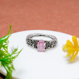 May include: A silver ring with a pink oval gemstone. The band features a floral design. The ring is set on a white surface with a yellow flower and green plant in the background.