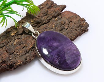 May include: Oval-shaped purple gemstone pendant with a silver-colored metal setting. The pendant is displayed on a piece of dark brown wood. Green leaves are visible in the upper left corner. The gemstone has a swirling pattern.