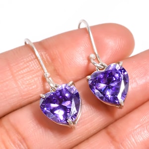 May include: A pair of heart-shaped purple gemstone earrings with silver hooks. The earrings are held in a hand, showcasing the vibrant colour and sparkle of the gemstones. The earrings are a romantic and elegant accessory.