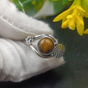 Puede incluir: Anillo de plata con una piedra de ojo de tigre pulida. El anillo presenta un diseño de hoja y flor. El ojo de tigre es de color marrón cálido con vetas doradas. Una joya.