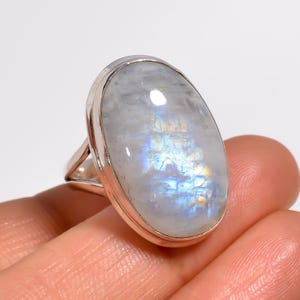 May include: A silver ring with a large, oval moonstone. The moonstone displays a milky white base with iridescent blue and yellow flashes. The ring's band is a simple, polished silver design.
