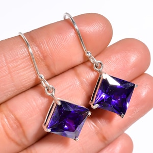 May include: Silver drop earrings with square, deep purple gemstones. The gemstones are set in a silver frame and hang from a simple hook. The earrings are shown against a light background.