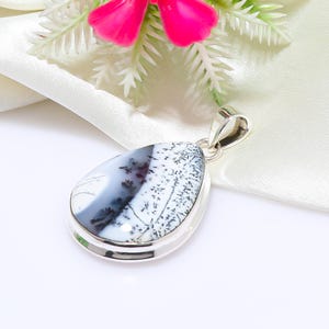 Peut inclure: Pendentif en argent en forme de goutte d'eau, orné d'une pierre d'agate dendritique. La pierre présente des inclusions noires et grises sur fond blanc. Le pendentif est présenté sur une surface blanche, avec des fleurs roses en arrière-plan.