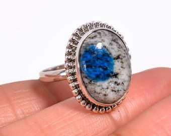 May include: Silver ring with an oval K2 stone. The stone has a white base with black flecks and a bright blue centre. The ring's bezel is decorated with small, beaded details, and the band is plain, polished silver.