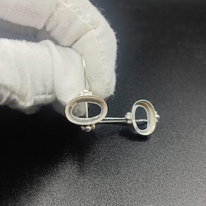 May include: A pair of silver earrings with an oval design and a small, round accent. The earrings are connected by a straight bar. The earrings are on a black surface.