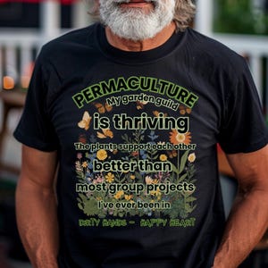 Puede incluir: Camiseta negra con el texto "PERMACULTURE My garden guild is thriving" en verde y blanco. La camiseta presenta un diseño floral con mariposas y la frase "DIRTY HANDS - HAPPY HEART".