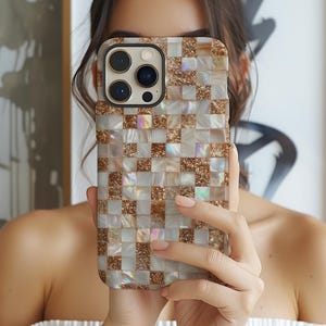May include: A phone case with a mosaic tile design. The case has a pattern of small, square tiles in white, gold, and iridescent colours. Some tiles have a glittery texture, adding sparkle. The phone case has a camera module with three lenses.
