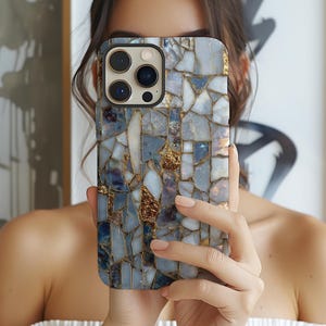 May include: A smartphone case with a mosaic design. The case features a pattern of irregular shapes in shades of blue, white, and gold. The camera lenses are visible at the top. The case is held in front of a person.