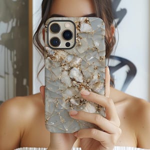 Puede incluir: Funda de teléfono inteligente con un patrón de piedra agrietada de color gris claro, acentuado con motas doradas y marrones. La funda tiene un borde negro y un recorte para la cámara. La funda se sostiene frente a la cara de una persona.