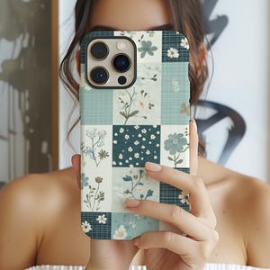 May include: A floral patterned phone case featuring a patchwork design with squares of teal, white, and navy blue. The case showcases various floral motifs, including daisies and other botanical elements. The case is designed for a smartphone.