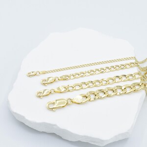 May include: Four gold chains of different thicknesses and lengths on a white surface. The chains are all made of a classic curb link style.