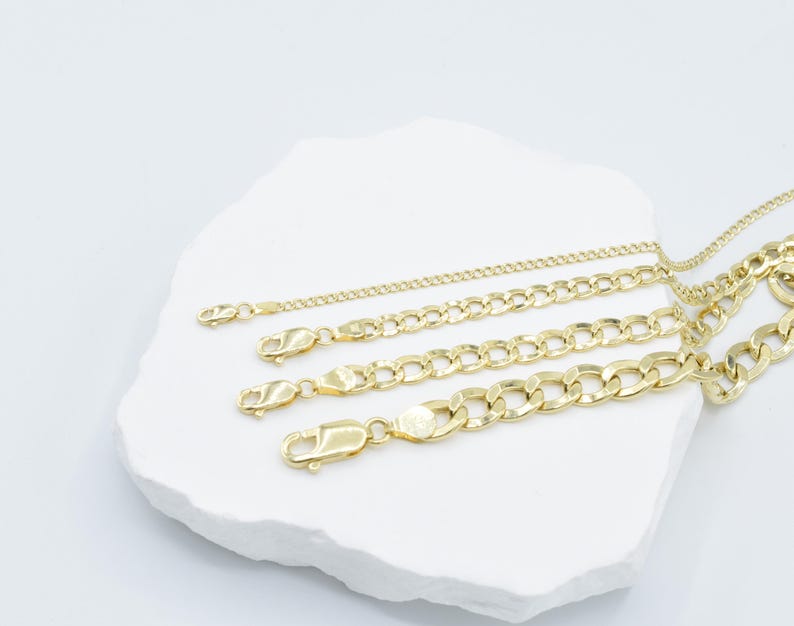 May include: Four gold chains of different thicknesses and lengths on a white surface. The chains are all made of a classic curb link style.