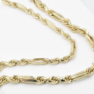 May include: A close-up shot of a gold-colored chain necklace. The necklace features a rope-like design with rectangular links. The chain is displayed against a white background, highlighting its shiny, reflective surface.