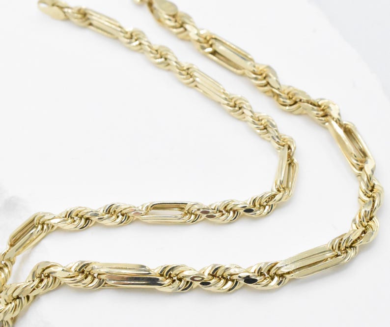 May include: A close-up shot of a gold-colored chain necklace. The necklace features a rope-like design with rectangular links. The chain is displayed against a white background, highlighting its shiny, reflective surface.