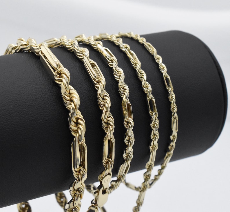 May include: Several gold-colored rope chains are displayed on a black cylindrical stand. The chains have a twisted rope design with rectangular links. The chains are made of a shiny, reflective material.