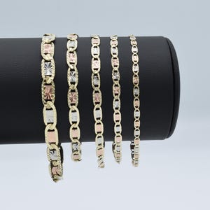 Könnte beinhalten: Fünf goldene Armbänder mit einem strukturierten, quadratischen Design. Die Armbänder werden auf einem schwarzen Ständer präsentiert.