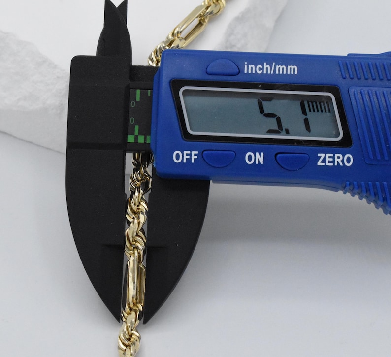 May include: A gold-colored rope chain is measured with a blue digital caliper. The caliper displays a measurement of 5.1 inches. The chain is placed between the caliper's jaws for an accurate reading. The caliper has an "OFF", "ON", and "ZERO" button.
