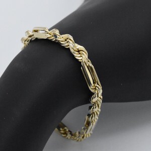May include: A gold-colored bracelet with a rope chain design and rectangular links. The bracelet is displayed on a black surface, showcasing its intricate details and polished finish. The jewelry piece is a classic accessory.