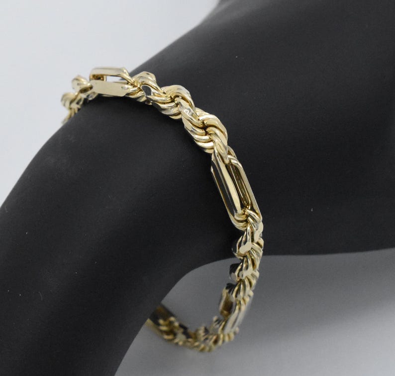 May include: A gold-colored bracelet with a rope chain design and rectangular links. The bracelet is displayed on a black surface, showcasing its intricate details and polished finish. The jewelry piece is a classic accessory.