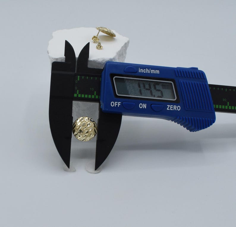 K&ouml;nnte beinhalten: Goldfarbene Ohrstecker mit strukturierter Oberfl&auml;che werden mit einem blauen digitalen Messschieber gemessen. Der Messschieber zeigt ein Ma&szlig; von 1,45 cm an. Zus&auml;tzliche Ohrringe befinden sich auf einer wei&szlig;en Oberfl&auml;che.