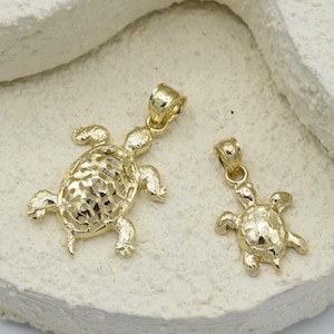 Könnte beinhalten: Zwei goldfarbene Schildkrötenanhänger. Der größere Anhänger hat ein durchbrochenes Design auf seinem Panzer. Der kleinere Anhänger hat einen soliden Panzer. Beide Anhänger haben eine Schlaufe für eine Kette.