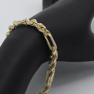 May include: A gold-colored bracelet with a rope chain design and rectangular links. The bracelet is made of a shiny, reflective metal and is displayed on a black surface. The jewelry piece is a classic accessory.