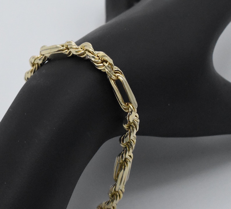 May include: A gold-colored bracelet with a rope chain design and rectangular links. The bracelet is made of a shiny, reflective metal and is displayed on a black surface. The jewelry piece is a classic accessory.