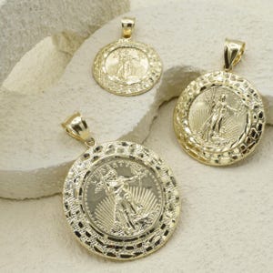 Puede incluir: Tres colgantes dorados con un diseño similar a una moneda. Los colgantes presentan una imagen central de una mujer sosteniendo una antorcha y una rama, rodeada por un borde texturizado. Los colgantes se exhiben sobre una superficie de color claro.