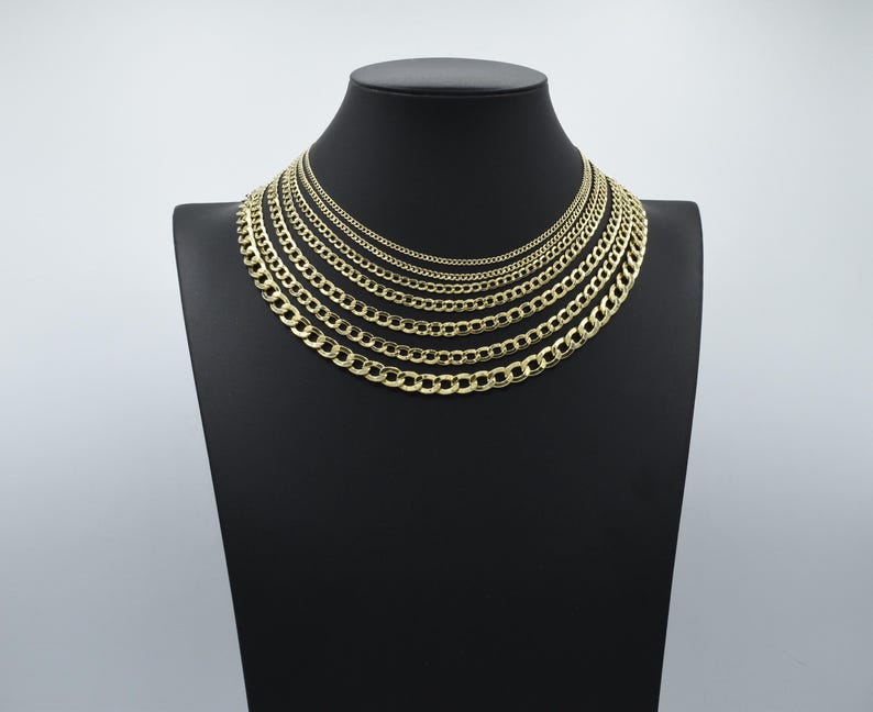 May include: A set of gold curb chain necklaces of varying widths displayed on a black velvet stand. The chains are arranged in order of size, with the smallest chain at the top and the largest chain at the bottom.