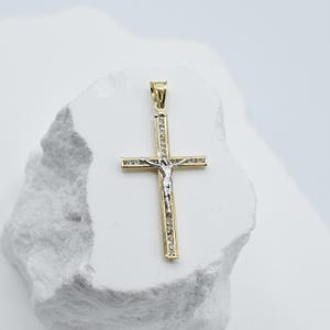 Op de afbeelding: Een gouden en zilveren kruis hanger met een klein figuurtje van Jezus Christus aan de voorkant. Het kruis is bezet met kleine, heldere stenen.