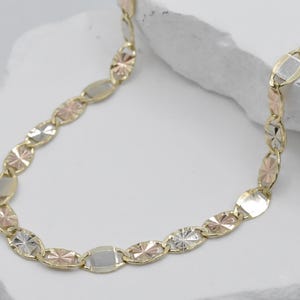 Puede incluir: Un collar de oro con eslabones ovalados y en forma de estrella en oro amarillo, rosa y blanco. La cadena se muestra sobre una superficie blanca, mostrando el dise&ntilde;o multicolor y los intrincados detalles de la joya.