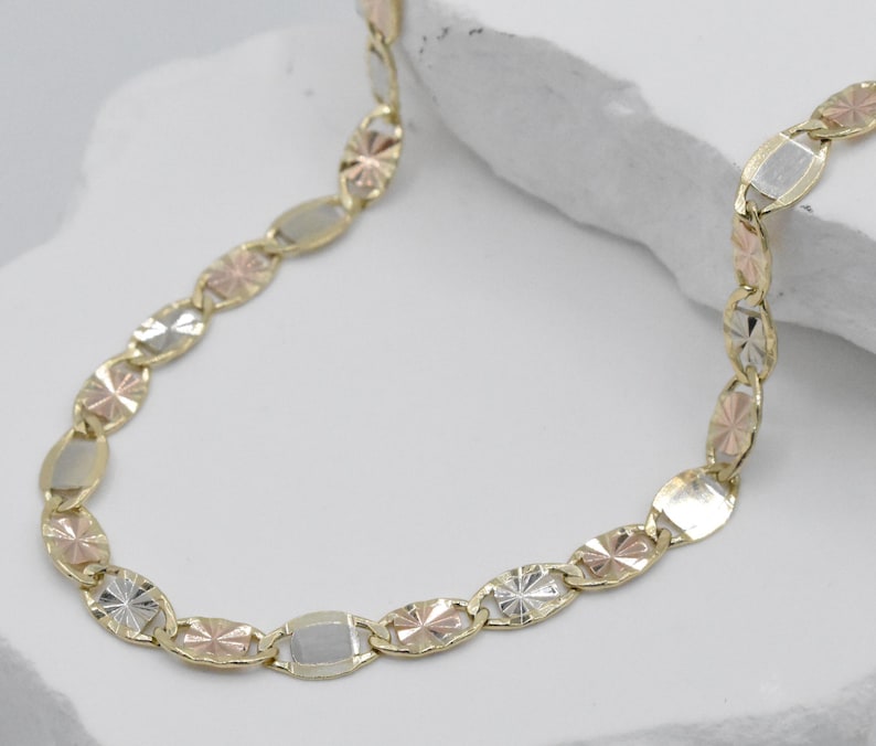 Puede incluir: Un collar de oro con eslabones ovalados y en forma de estrella en oro amarillo, rosa y blanco. La cadena se muestra sobre una superficie blanca, mostrando el dise&ntilde;o multicolor y los intrincados detalles de la joya.
