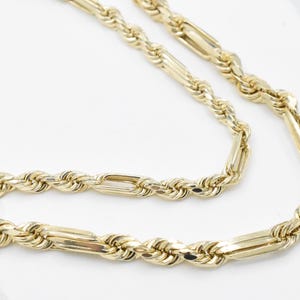 May include: A gold-colored necklace with a rope chain design and rectangular links. The necklace is displayed on a white surface, showcasing its intricate details and polished finish. The chain is made of metal.