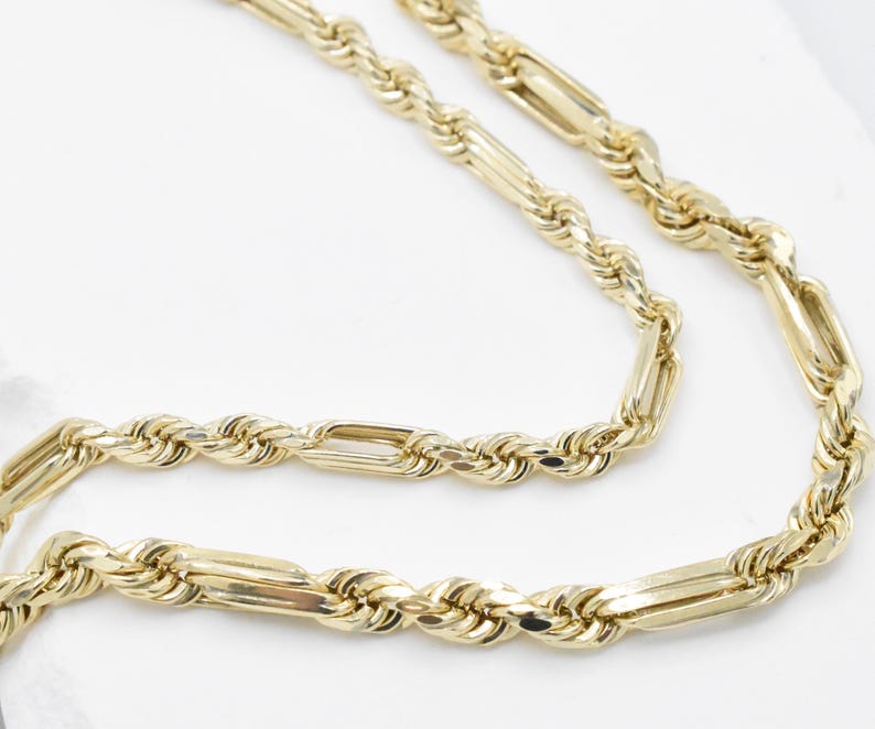 May include: A gold-colored necklace with a rope chain design and rectangular links. The necklace is displayed on a white surface, showcasing its intricate details and polished finish. The chain is made of metal.