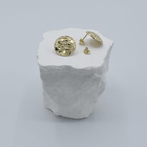 K&ouml;nnte beinhalten: Goldfarbene Ohrstecker mit strukturiertem, durchbrochenem Design. Ein Ohrring ist rund, der andere hat die Form einer flachen Scheibe. Ein Paar Ohrringverschl&uuml;sse sind ebenfalls sichtbar. Die Ohrringe werden auf einer wei&szlig;en, strukturierten Oberfl&auml;che pr&auml;sentiert.