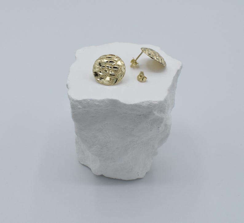 K&ouml;nnte beinhalten: Goldfarbene Ohrstecker mit strukturiertem, durchbrochenem Design. Ein Ohrring ist rund, der andere hat die Form einer flachen Scheibe. Ein Paar Ohrringverschl&uuml;sse sind ebenfalls sichtbar. Die Ohrringe werden auf einer wei&szlig;en, strukturierten Oberfl&auml;che pr&auml;sentiert.