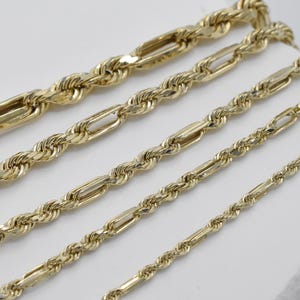 May include: Close-up of several gold-colored chain necklaces. The chains vary in style, including rope and Figaro designs. The chains are displayed on a white surface, showcasing their intricate details and polished finish. The chains are of different lengths.