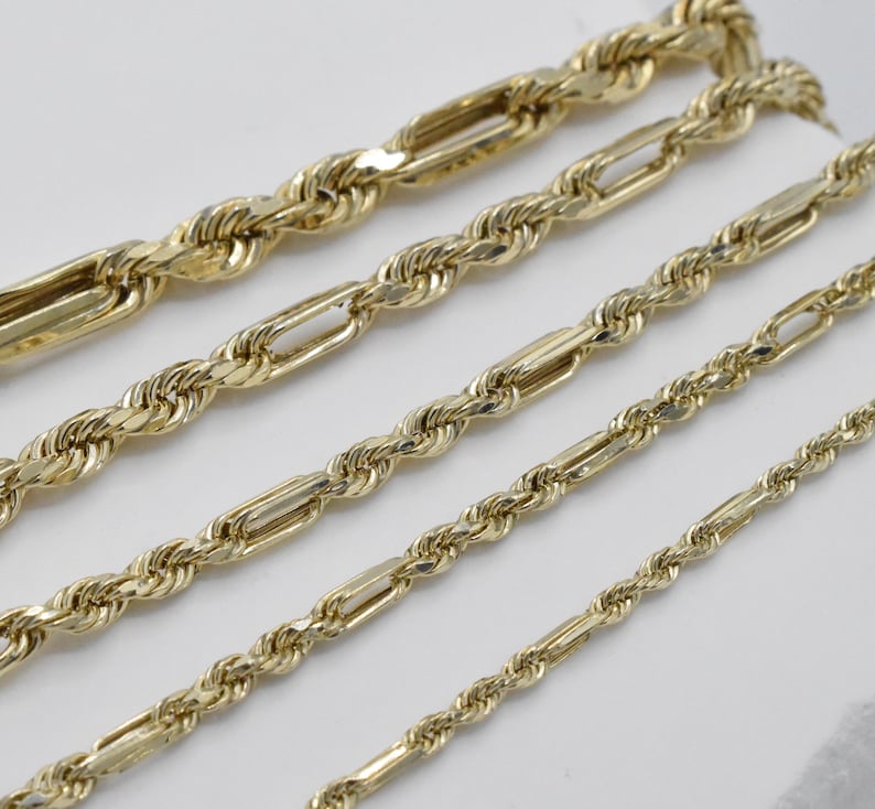 May include: Close-up of several gold-colored chain necklaces. The chains vary in style, including rope and Figaro designs. The chains are displayed on a white surface, showcasing their intricate details and polished finish. The chains are of different lengths.