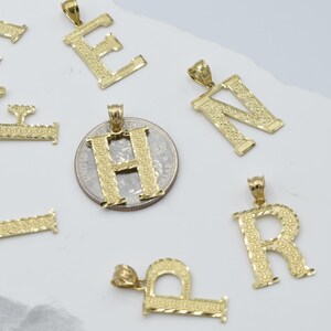 Puede incluir: Un conjunto de colgantes de letras del alfabeto chapados en oro. Las letras son A, E, F, H, N, P y R. Los colgantes est&aacute;n sobre una superficie blanca.