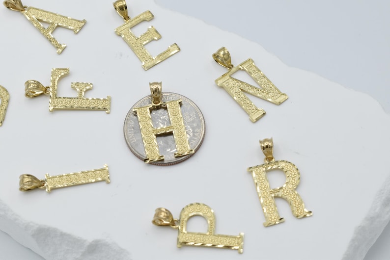 Puede incluir: Un conjunto de colgantes de letras del alfabeto chapados en oro. Las letras son A, E, F, H, N, P y R. Los colgantes est&aacute;n sobre una superficie blanca.