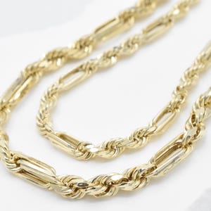 May include: A gold-colored necklace with a Figaro chain design. The necklace features alternating elongated rectangular links and twisted rope-like links. The jewelry is displayed on a white surface.