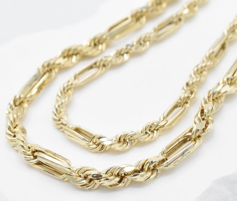 May include: A gold-colored necklace with a Figaro chain design. The necklace features alternating elongated rectangular links and twisted rope-like links. The jewelry is displayed on a white surface.