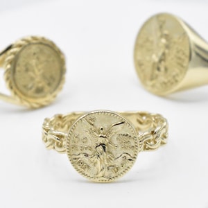 May include: Three gold rings with coin-like designs. One ring has a heart-shaped red stone. The rings are made of gold and feature detailed coin engravings. The rings are displayed on a white surface.
