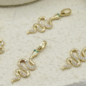 Puede incluir: Colgantes dorados con forma de serpiente con detalles blancos y verdes. Los amuletos con forma de serpiente están adornados con pequeñas piedras blancas brillantes a lo largo de sus cuerpos y tienen ojos verdes. Los colgantes se exhiben sobre una superficie clara.