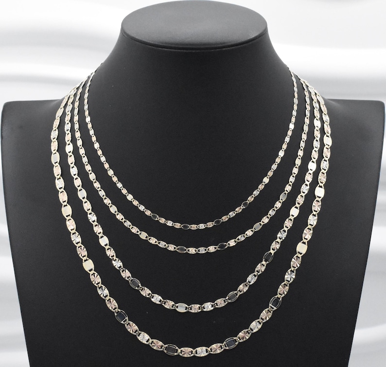 Puede incluir: Conjunto de collares plateados expuestos sobre un maniqu&iacute; negro. Los collares, de diferentes longitudes, presentan cada uno una delicada cadena con eslabones ovalados. Las cadenas est&aacute;n dispuestas en capas, creando un efecto en cascada.