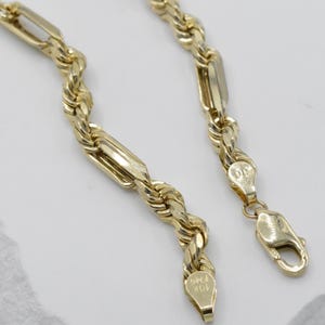 May include: A gold-colored necklace with a rope chain design, featuring rectangular links and a lobster clasp. The necklace is made of 10K gold and is displayed on a light-colored surface.