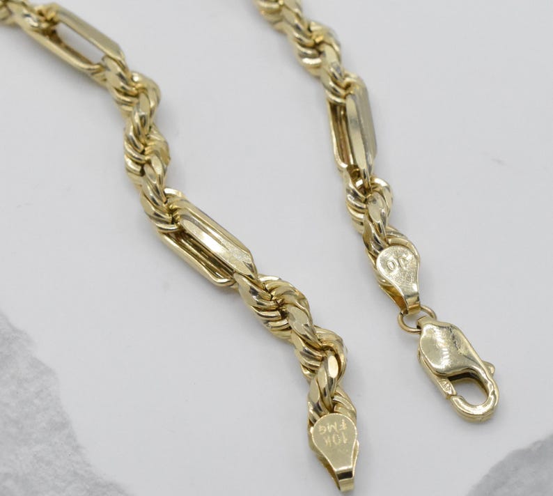 May include: A gold-colored necklace with a rope chain design, featuring rectangular links and a lobster clasp. The necklace is made of 10K gold and is displayed on a light-colored surface.