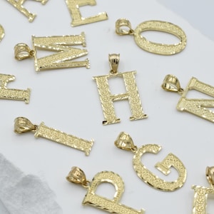 Puede incluir: Una colecci&oacute;n de colgantes de alfabeto de tono dorado con un acabado texturizado. Los colgantes est&aacute;n dispuestos sobre una superficie blanca.