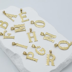 Puede incluir: Una colecci&oacute;n de colgantes de letras del alfabeto chapados en oro. Cada letra est&aacute; dise&ntilde;ada de forma intrincada con una superficie texturizada.